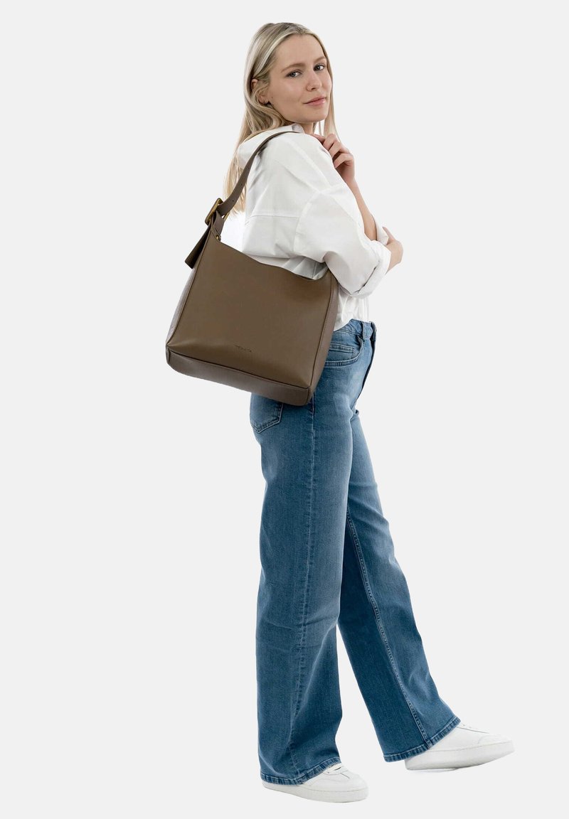 Donna che indossa una camicia bianca, jeans blu, sneakers bianche e che porta una grande borsa a tracolla marrone sulla spalla destra, su uno sfondo semplice.