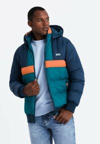 Fodrad jacka i marinblått, teal och orange med huva. Har dragkedjestängning, elastiska ärmslut och en logotypapplikation på bröstet.