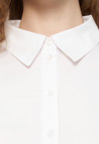 Λευκό πουκάμισο με κουμπιά, κλασικό γιακά, απαλό υφασμά, μικρά στρογγυλά κουμπιά και διακριτική γυαλάδα. Κοντινή λήψη του γιακά και της σειράς με τα κουμπιά.