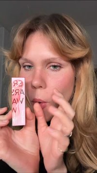 Lipstick in a pink rectangular tube with glossy finish. Label displays product name in bold letters. Model holds tube close to face.