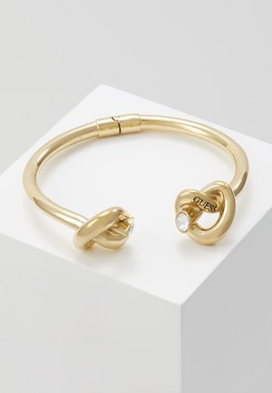 Bracelet jonc ouvert doré avec deux extrémités en forme de cœur entrelacées, chacune ornée d'un cristal clair et un des cœurs portant le logo "GUESS".