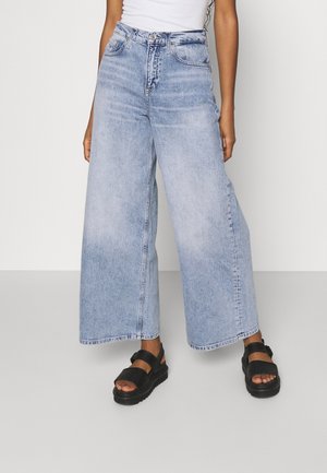 Personne portant un jean bleu clair à jambes larges, des sandales compensées noires et un haut blanc sans manches sur un fond clair uni.