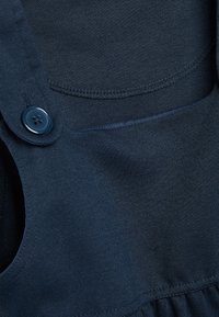 Granatowy materiał o gładkiej fakturze, z zapięciem na guzik i wszytą kieszonką. Szwy są starannie wykończone.