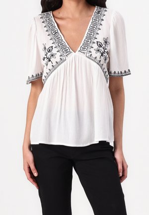 White blouse with black floral embroidery along deep V-neckline, sleeves, and chest, paired with black pants on a standing person.