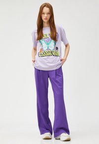 Camiseta gráfica lila con estampado de mariposa, combinada con pantalones morados de corte amplio. Usada con zapatillas deportivas blancas.
