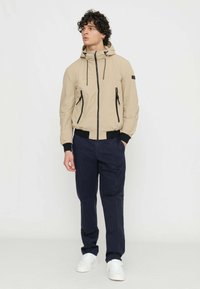 Giacca beige con cappuccio e dettagli neri, chiusura frontale con zip e coulisse, abbinata a pantaloni blu scuro e scarpe da tennis bianche. Tessuto liscio.