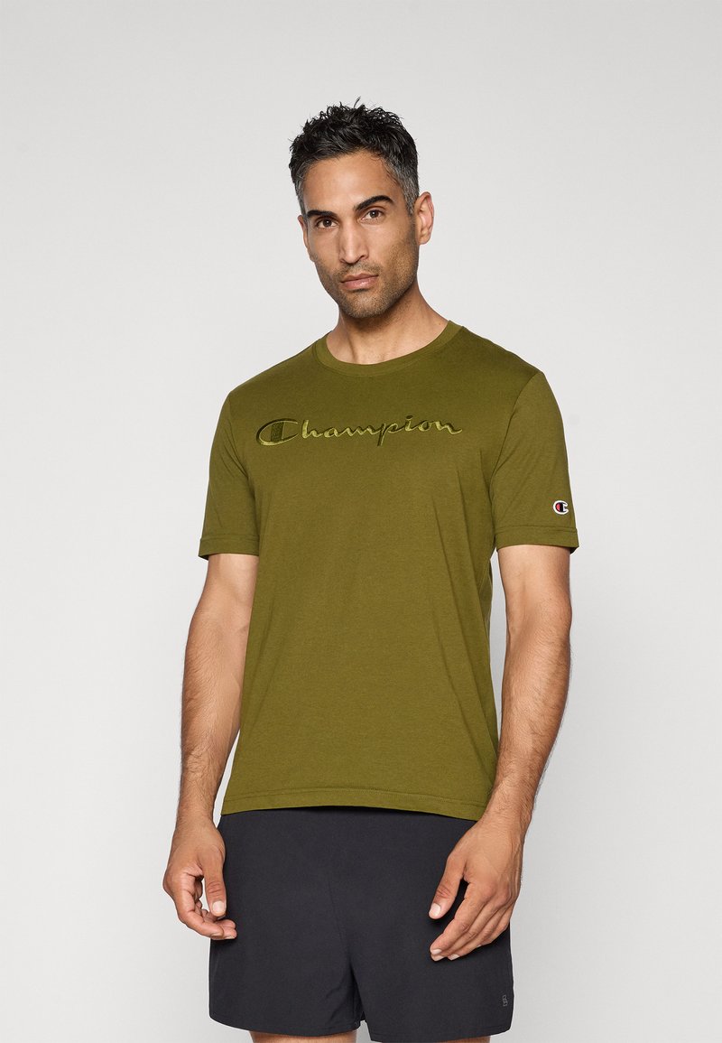 Mann mit einem olivgrünen Champion-T-Shirt mit gesticktem Logo und schwarzen Shorts, der vor einem schlichten hellen Hintergrund steht.