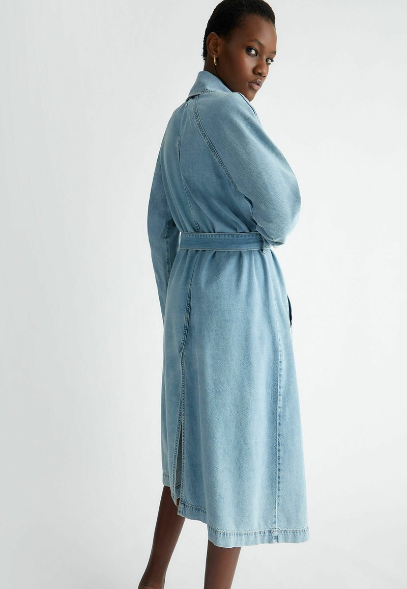 Cappotto trench in denim blu chiaro con vita regolabile, maniche lunghe e spacchi laterali. Presenta un colletto e dettagli di cucitura in vista.