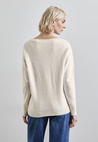 Donna con capelli biondi corti, che indossa un maglione crema a maniche lunghe e jeans blu, di spalle, di fronte a una parete grigio chiaro.