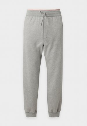 Mand iført sort polo med rød krave og manchetter, lysegrå Versace sweatpants, hvide og brune sneakers og solbriller, stående med den ene hånd i lommen.