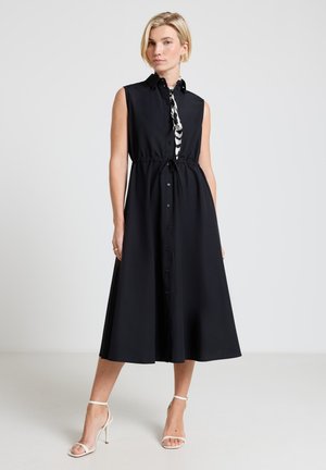 Woman stands wearing sleeveless black button-down midi dress with waist tie, paired with white high-heeled sandals and a black-and-white neck scarf.