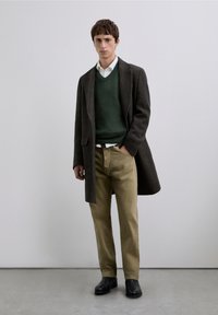 Abrigo de lana gris sobre un suéter verde, camisa blanca y pantalones beige. El modelo lleva zapatos negros. Diseño sencillo, colores neutros.