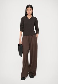 Ruskea neulottu V-pääntie villapaita pitkillä hihoilla, yhdistettynä leveisiin ruskeisiin housuihin. Malli pitää mustaa laukkua kädessään seisten valkoista taustaa vasten.