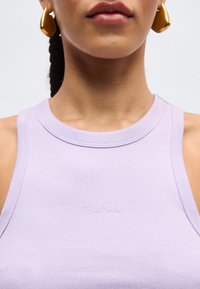 Κοντινό πλάνο ενός ατόμου που φοράει ένα λιλά ριμπ μπλουζάκι χωρίς μανίκια με μικρό κεντητό κείμενο και μεγάλα χρυσά σκουλαρίκια σε σχήμα σταγόνας.