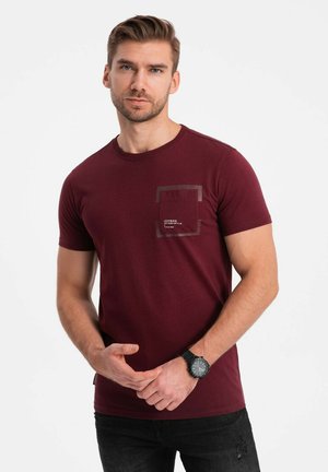 Burgundy short-sleeve t-shirt with textured fabric, a square pocket with graphic detail, and a round neckline. Worn with dark jeans.