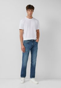 T-shirt blanche à manches courtes avec un col rond, associée à un jean en denim bleu. Le modèle porte des baskets blanches ; le décor est minimaliste.
