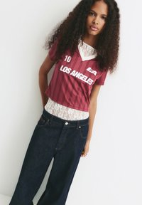 Ung kvinde med langt krøllet hår iført en bordeaux "Los Angeles" sports t-shirt over en hvid blonde top og mørke brede jeans, poserer mod en hvid væg.