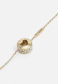 Bracelet en or avec un charme circulaire comportant une découpe en forme de cœur et des cristaux transparents, gravé avec le mot "Guess" et accentué par une chaîne.