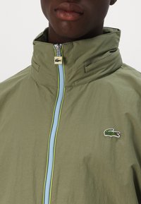 Olivovo zelená bunda má vysoký golier a predný zips s modrými akcentmi. Vyšívané logo Lacoste sa nachádza na ľavej strane. Hladký povrch.