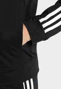 Main dans la poche d'un survêtement noir Adidas avec un design à trois bandes blanches sur la manche et le pantalon, sur un fond clair uni.
