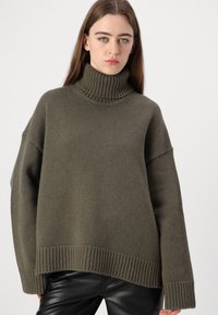 Overdimensjonert genser med høy hals i mørk olivengrønn strikket stoff. Har ribbestrikket krage og kant, drop shoulders, og en strukturert overflate.