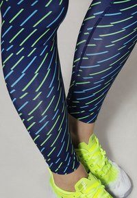 Leggings blu navy con linee diagonali verdi e blu, realizzati in un materiale elasticizzato. Abbinati a scarpe da ginnastica giallo brillante.