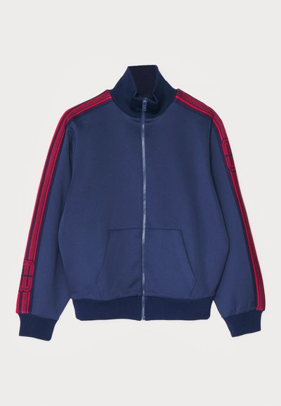 Giacca a track in tinta blu navy con zip e colletto alto, dettagli a strisce rosse sulle maniche e tasche frontali. Tessuto morbido con polsini a costine.