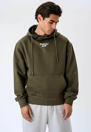Homme portant un sweat à capuche vert olive avec le texte « Toujours Actif », regardant vers le bas, associé à un pantalon gris clair sur fond uni.