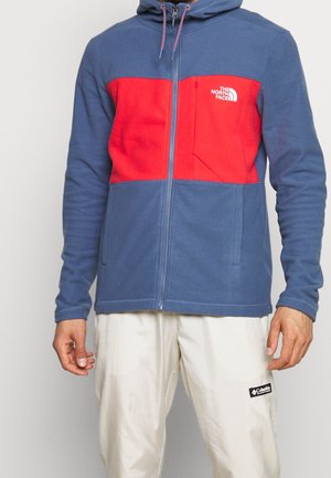 Veste polaire zippée bleue et rouge avec capuche, dotée d'une poche avant et d'un logo sur la poitrine. Portée avec un pantalon de couleur crème.
