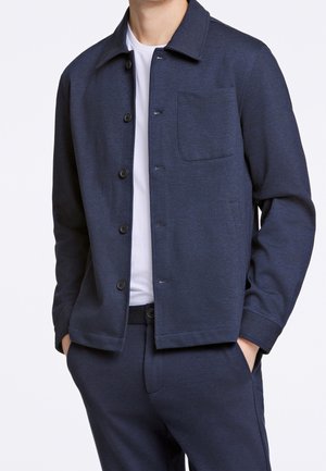 Mand iført navyblå jakke med knapper og brystlomme over hvid T-shirt, matchende navybukser, hænder i lommer, foran hvid baggrund.