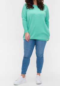 Žena stojící, oblečená v volném světle zeleném svetru, modrých úzkých džínách a bílých teniskách, na jednobarevném bílém pozadí.