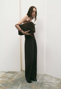 Bolso de ante marrón con forma rectangular, sujetado bajo el brazo; combinado con pantalones negros holgados y una blusa con encaje en los bordes. Fondo neutro.