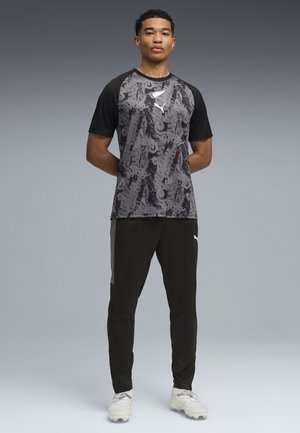 Jonge man staat met handen achter de rug, draagt een zwart-grijs sportshirt met patroon, zwarte broek en witte voetbalschoenen tegen een grijze achtergrond.