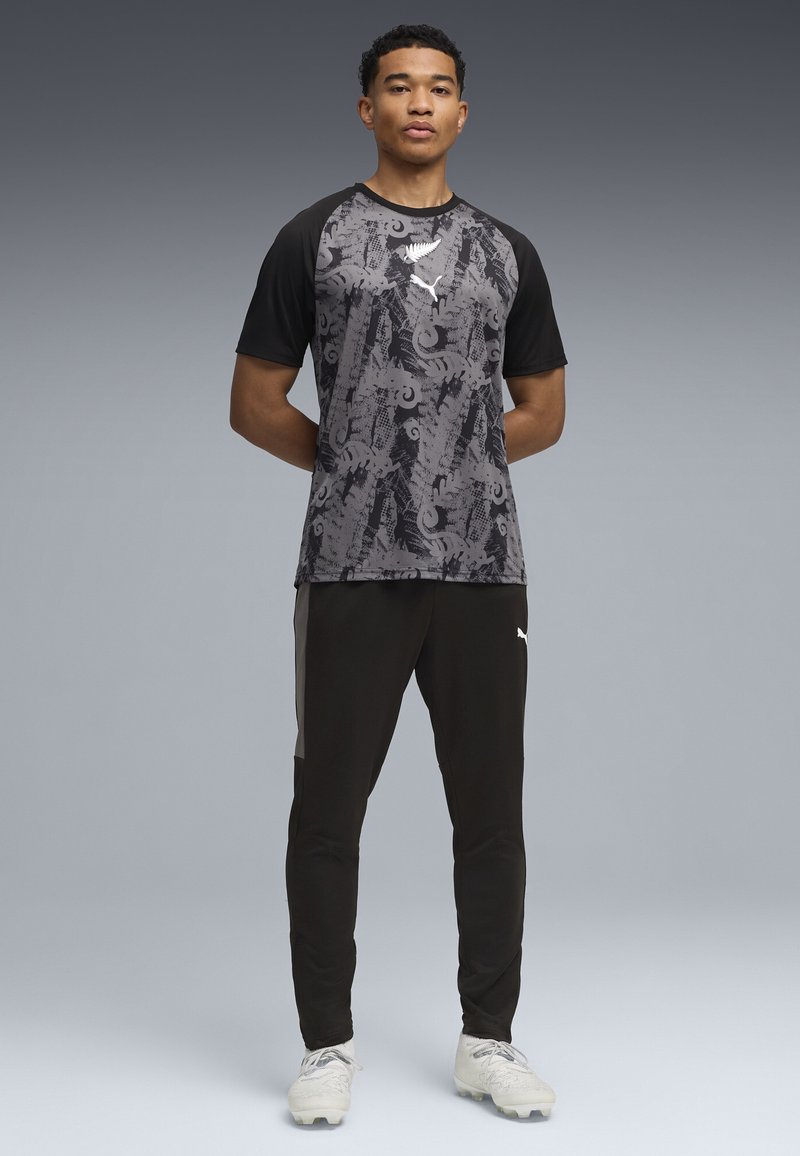 Jeune homme debout, les mains derrière le dos, portant un maillot de sport noir et gris à motifs, un pantalon noir et des crampons blancs, sur un fond gris.
