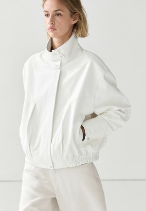Vrouw met kort blond haar, die een witte jas draagt met een hoge kraag, drukknopen en een elastische zoom, staand met handen in de zakken tegen een effen achtergrond.