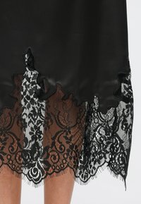 Zwarte satijnen jurk met een gedetailleerde zwarte kanten zoom met bloemmotieven en een geschulpte rand. De stof is glad met een lichte glans.