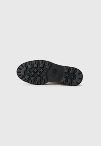 Semelle de botte noire avec un motif de semelle robuste pour l'adhérence, présentant des crampons en forme de croix et rectangulaires sur fond blanc uni.