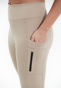 Leggings de sport beiges avec une taille haute, poche latérale avec une fermeture éclair noire et des détails de couture texturés. Tissu lisse et extensible.