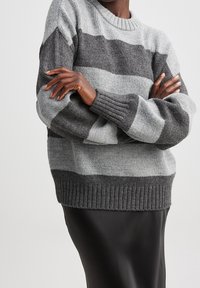 Harmaa ja vaaleanharmaa raidoitettu neulepaita, jossa on pyöreä pääntie ja ribattuja hihansuita, päällä mustan hameen kanssa.