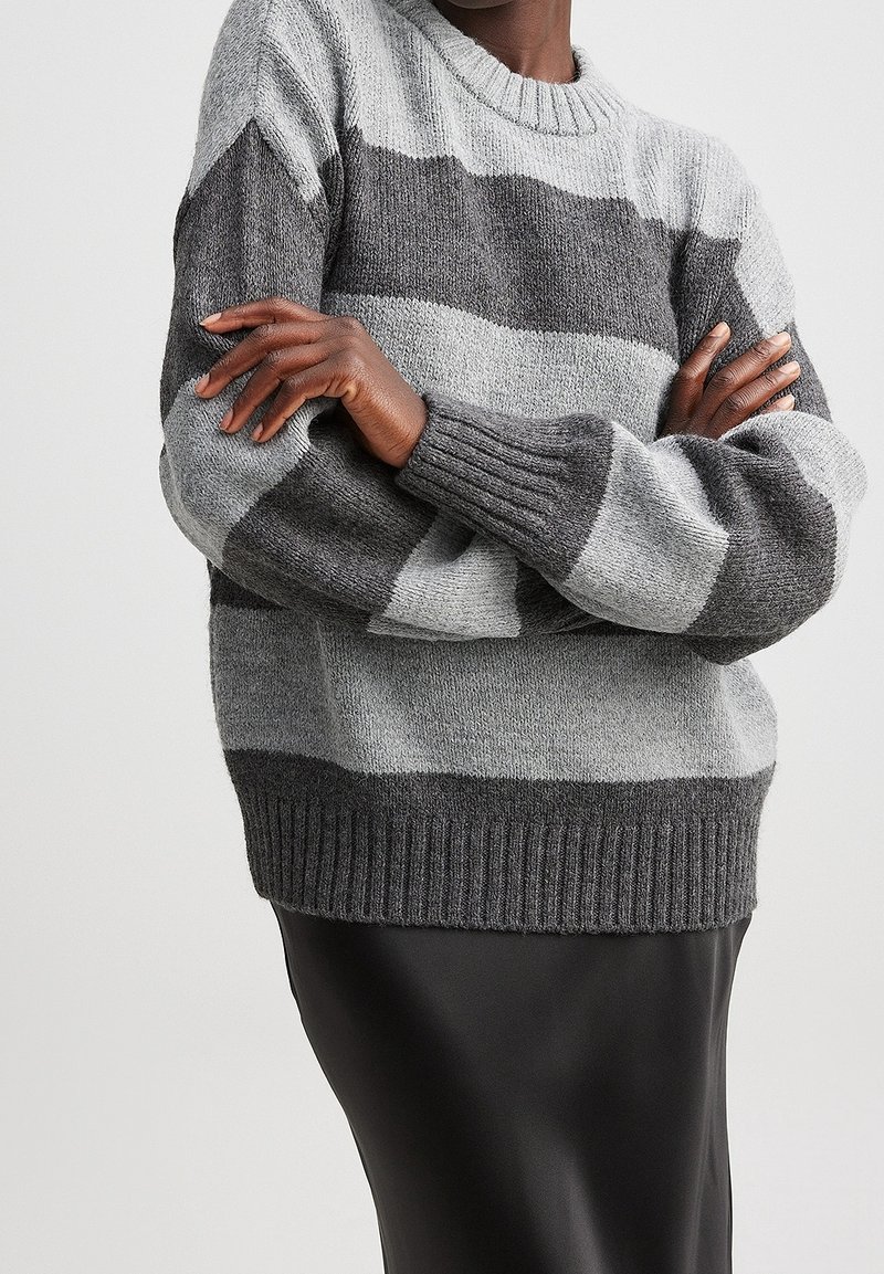 Harmaa ja vaaleanharmaa raidoitettu neulepaita, jossa on pyöreä pääntie ja ribattuja hihansuita, päällä mustan hameen kanssa.