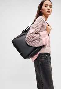 Geantă tote din piele neagră cu formă dreptunghiulară elegantă, prevăzută cu mânere robuste. Purtată peste un pulover cu dungi roz și gri și o blugi închiși la culoare.