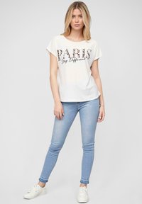 T-shirt blanc avec "PARIS" en imprimé animal et "Stay Different" en écriture noire, associé à un jean skinny bleu clair et des baskets blanches.