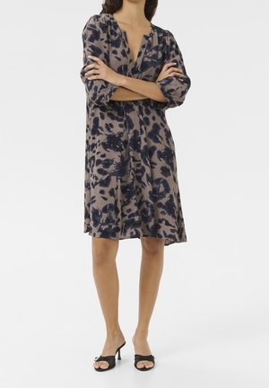 Woman wearing a knee-length, long-sleeve dress with abstract navy and beige pattern, standing with arms crossed and black heeled sandals.