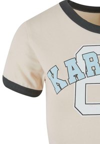 Bežs kokvilnas T-krekls ar apaļu melnu ribota kakla izgriezumu, priekšpusē ar gaiši zilu grafisko tekstu "KARL" un skaitli "8".