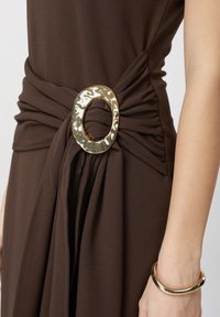 Robe marron foncé avec tissu froncé et une grande boucle ovale en or martelé à la taille, portée avec un bracelet épais en or au poignet.