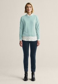 Ljusblå tröja av mjuk textil, lager på lager över en vit skjorta; matchad med mörkblå byxor och svarta ankelstövlar.