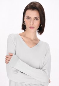 Hellgrauer Strickpullover mit V-Ausschnitt und langen Ärmeln. Mit gerippten Bündchen und einer soften Textur, getragen mit über dem Kreuz gelegten Armen.