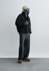 Černá fleecová bunda, šedé cargo kalhoty, hnědé nazouvací boty a béžová helma. Model stojí v profilu na jednoduchém pozadí.