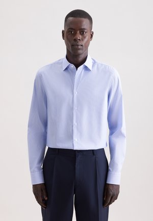 Man die staat en een lichtblauw overhemd met lange mouwen draagt, ingestopt in donkerblauwe broek, tegen een witte achtergrond.