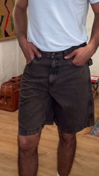 Άτομο που φοράει λευκό t-shirt και μαύρο τζιν σορτς με τα χέρια στις τσέπες, στέκεται σε εσωτερικό χώρο σε ξύλινο πάτωμα.